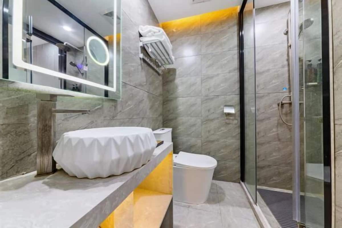 Un baño moderno con suelo de mármol, cabina de ducha de cristal, lavabo blanco con encimera de mármol y espejo con luz circular.