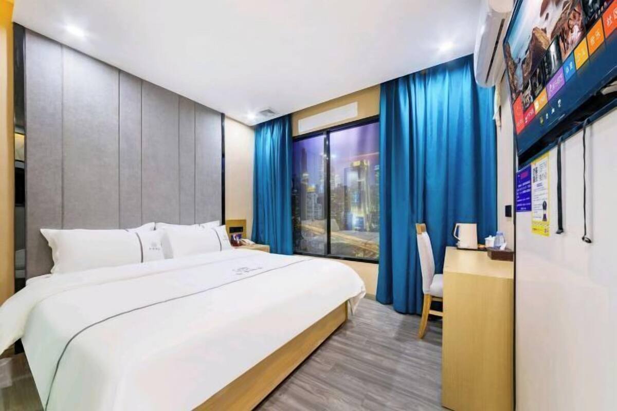 Habitación de hotel con una cama grande, un escritorio, una silla, un ventanal con cortinas y un televisor montado en la pared.