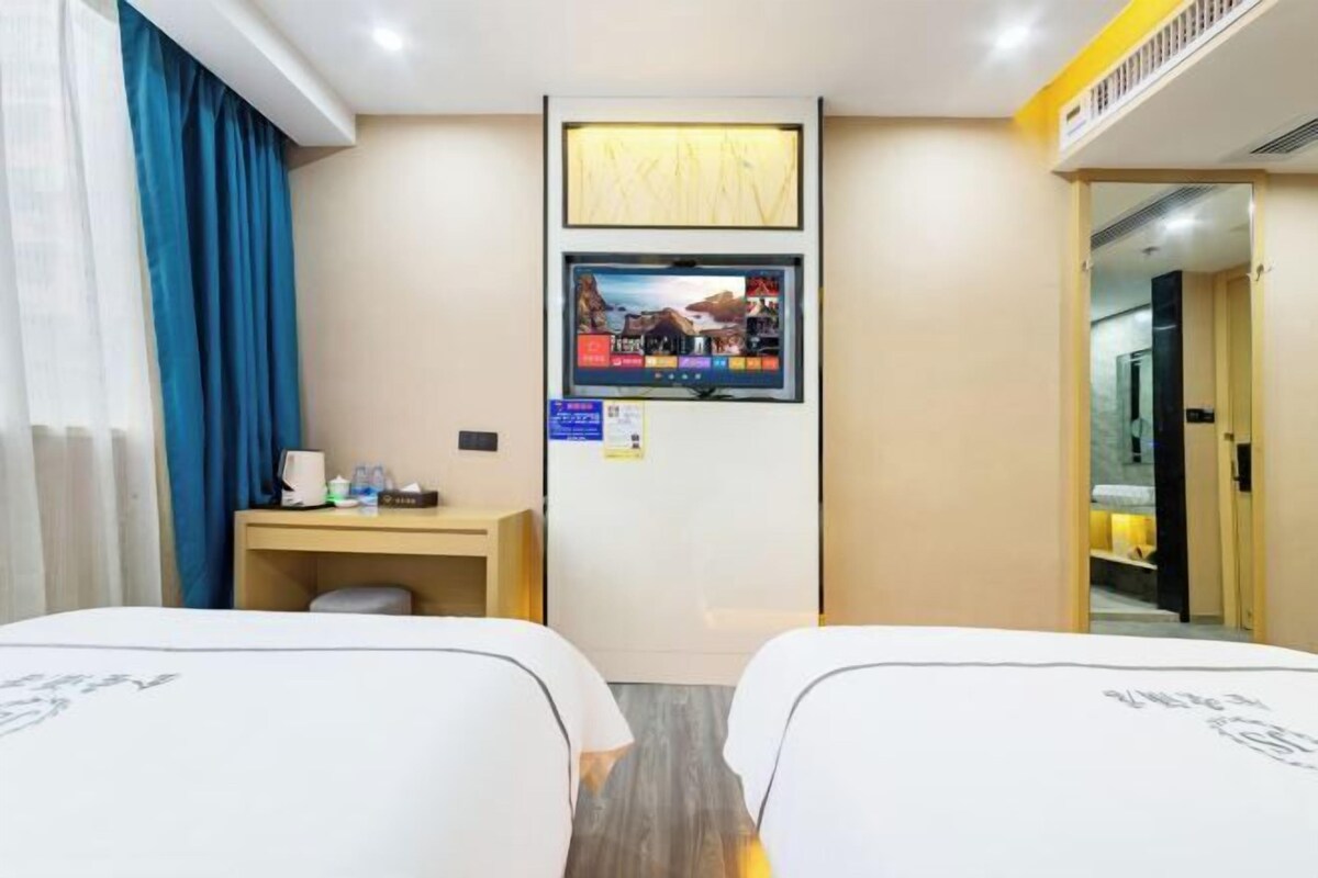 Hotellihuone, jossa on kaksi sänkyä, työpöytä tietokoneella, seinälle asennettu televisio ja ovi, joka johtaa toiseen huoneeseen.