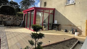 Un espace patio doté d’une terrasse en bois, d’un petit arbre et d’une véranda en verre.