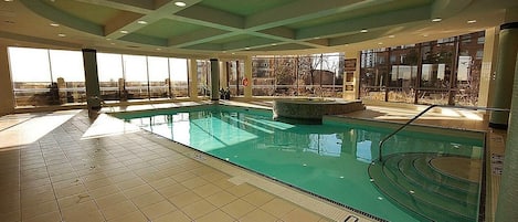 Une piscine intérieure à l’eau claire, dotée d’un sol carrelé où sont indiquées les profondeurs, et de grandes fenêtres offrant une vue sur l’extérieur.