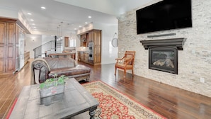 A spacious living room with a fireplace, a large TV mounted on a stone wall, and a kitchen in the background. - The Castle-Pool/Spa/Billiard (Henderson)