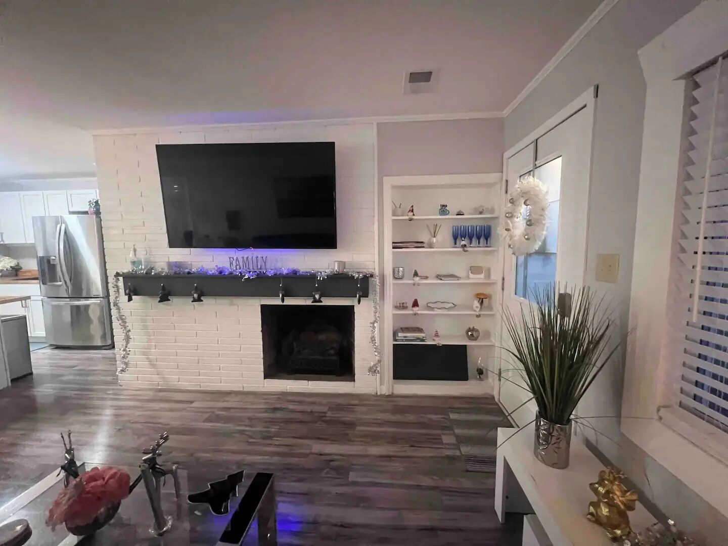 Ein modernes Wohnzimmer mit Flachbildfernseher über einem Kamin, einer Küchenzeile mit Edelstahlgeräten und einem dekorativen Regal mit verschiedenen Gegenständen.