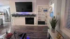Ein modernes Wohnzimmer mit Flachbildfernseher über einem Kamin, einer Küchenzeile mit Edelstahlgeräten und einem dekorativen Regal mit verschiedenen Gegenständen.