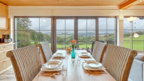 A dining table set for four with wicker chairs, large windows offering a view of a green landscape, and a kitchen area with wooden cabinets and a coffee maker.
