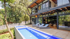 Una casa moderna a due piani con una grande piscina, area salotto all'aperto e vista sulla foresta circostante.