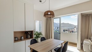 A modern dining area with a wooden table and black chairs, a hanging light fixture, and a view of buildings and mountains through a glass door. - Harbor & Sea View Agaete (Puerto de las Nieves)