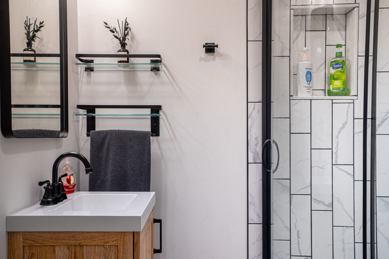 Un baño moderno con un lavamanos blanco, un grifo negro y una toalla colgada en un perchero. Hay dos pequeñas macetas con plantas en un estante sobre el lavamanos.
