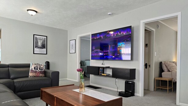 Een moderne woonkamer met een grote flatscreen televisie aan de muur, een houten salontafel, een zwarte leren bank en een klein bijzettafeltje met een bloevas.