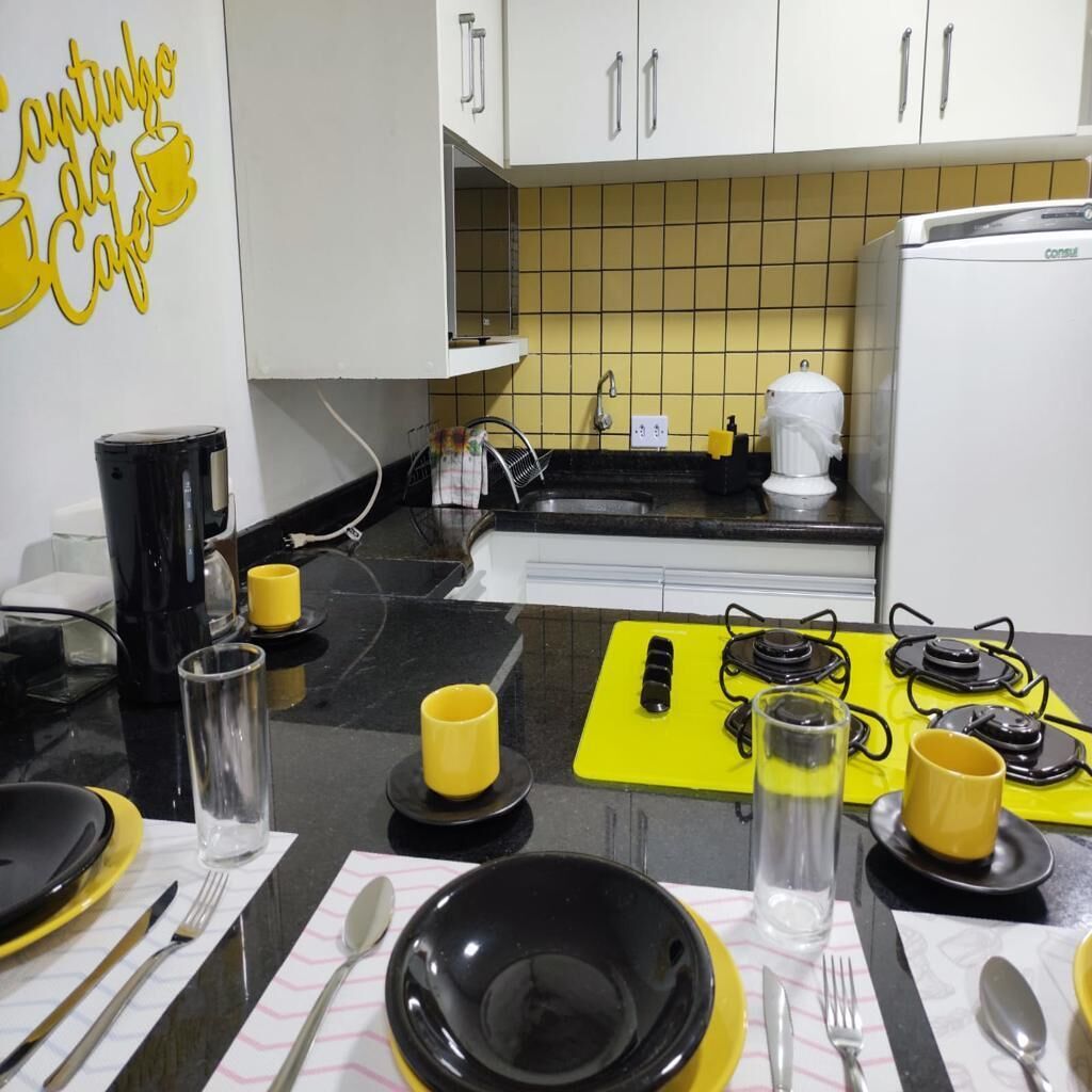 Een keuken met witte kasten, een geel betegelde spatwand, een zwart aanrechtblad en een gele placemat met bestek.