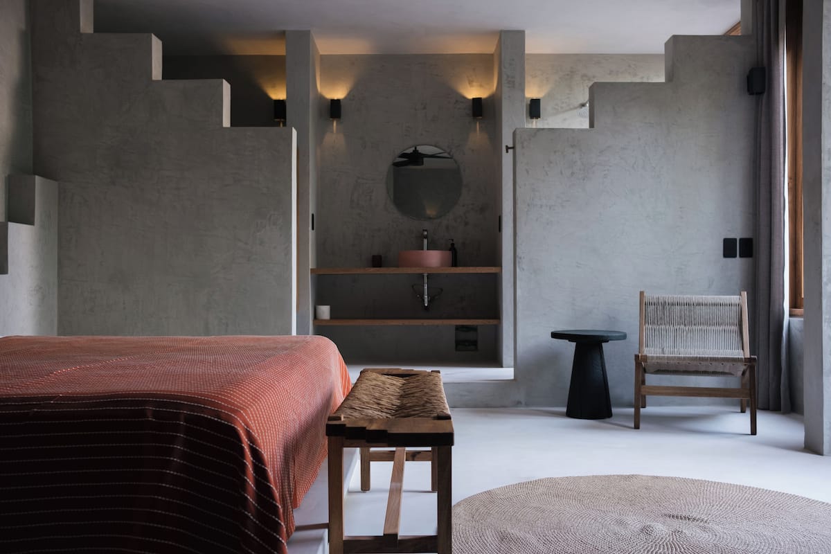 A modern bedroom with a bed, a wooden bench, a round table, a chair, and a built-in sink with a mirror.