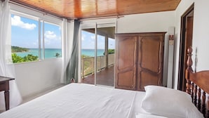 A bedroom with a large bed, a wooden wardrobe, and a balcony with a view of the ocean.