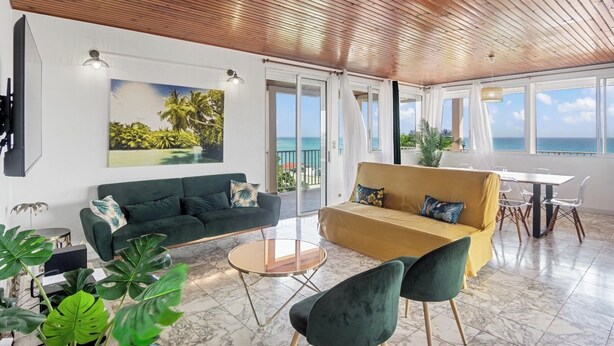 A spacious living area with a green sofa, a yellow sofa, and a coffee table. There is a large window with a view of the ocean.
