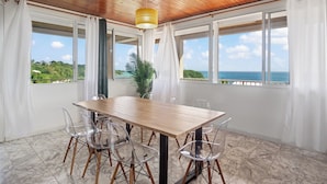 A dining area with a wooden table and chairs, large windows with curtains, a hanging light fixture, and a view of the ocean.