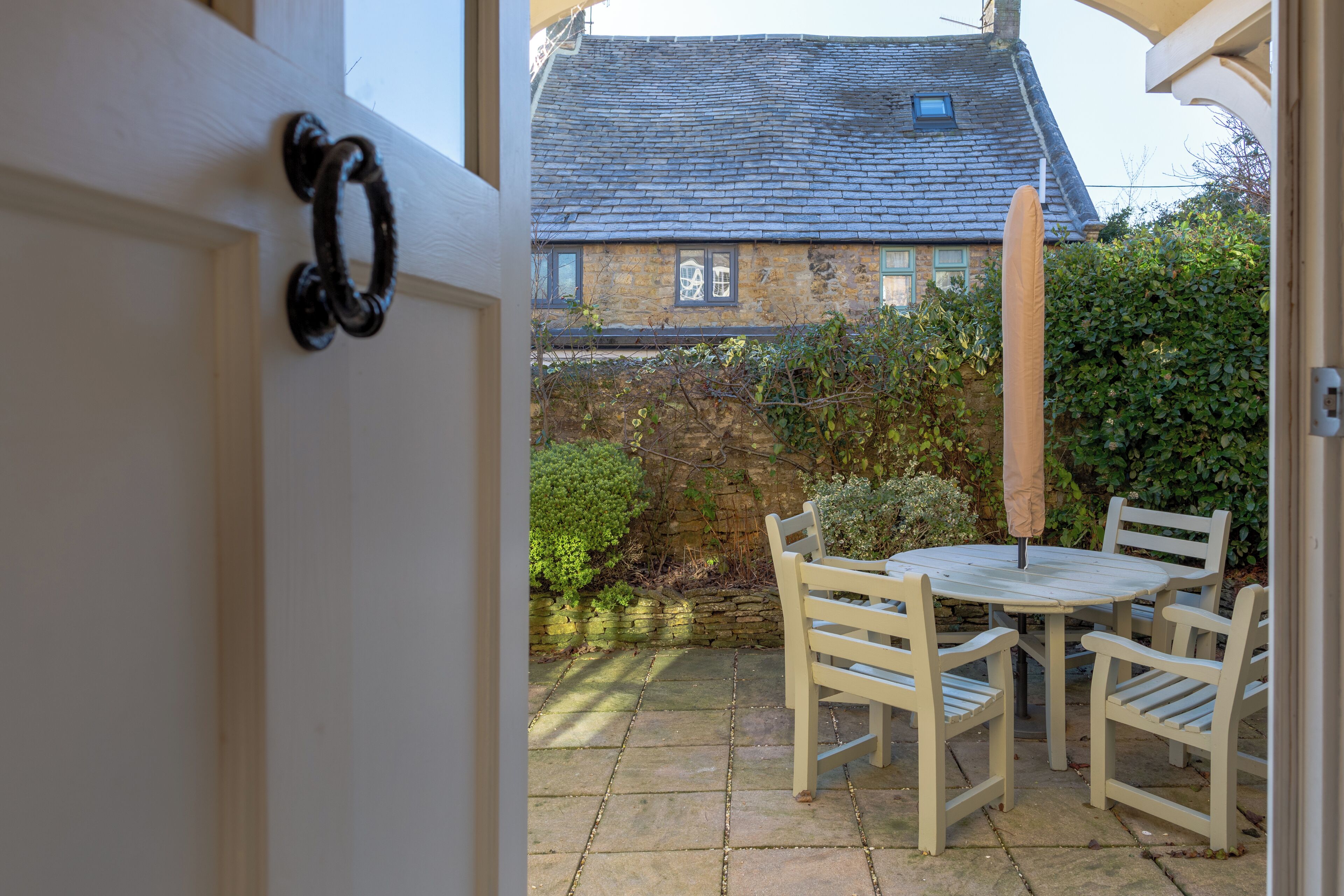 A stone patio with a round table and four chairs, a wooden door with a handle, and a stone wall with climbing plants.