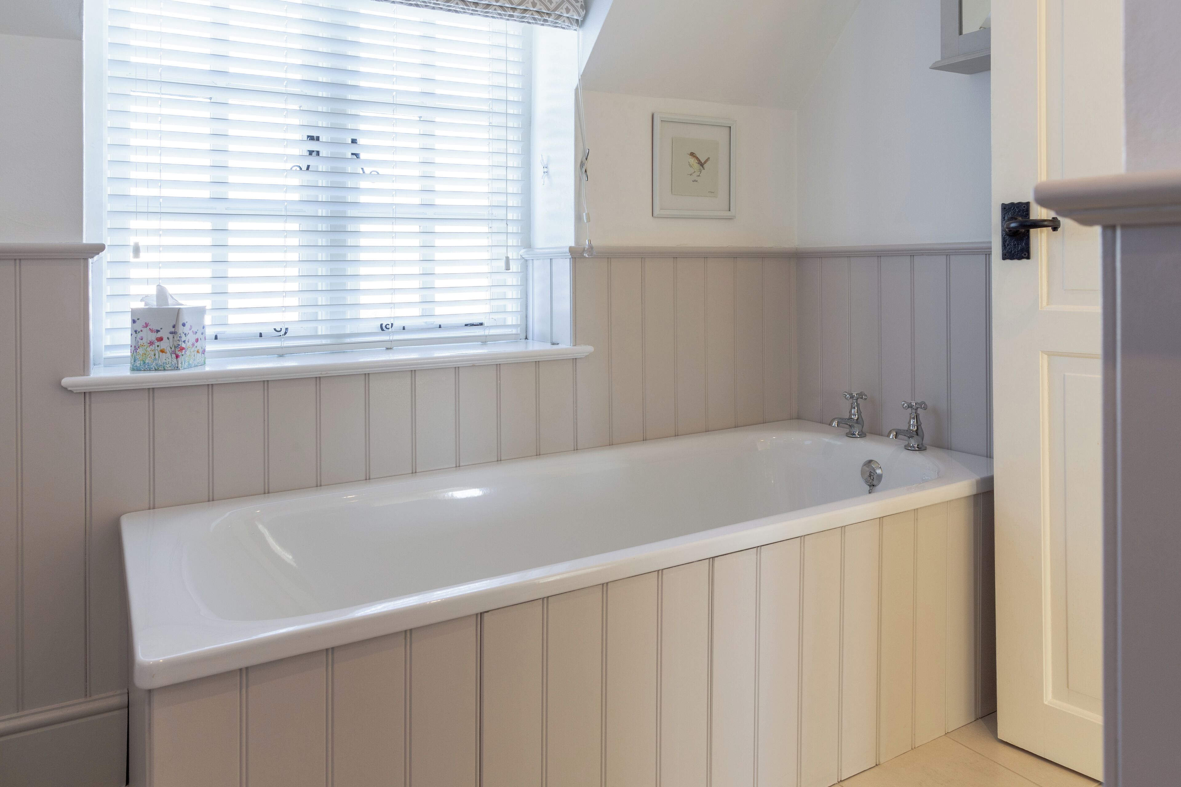 A bathroom with a white bathtub, a window with blinds, and a framed picture on the wall.