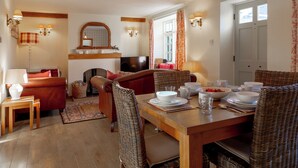 Un comedor con una mesa de madera dispuesta para cuatro personas, rodeada de sillas de mimbre. En el fondo, un sofá rojo y un televisor son visibles.