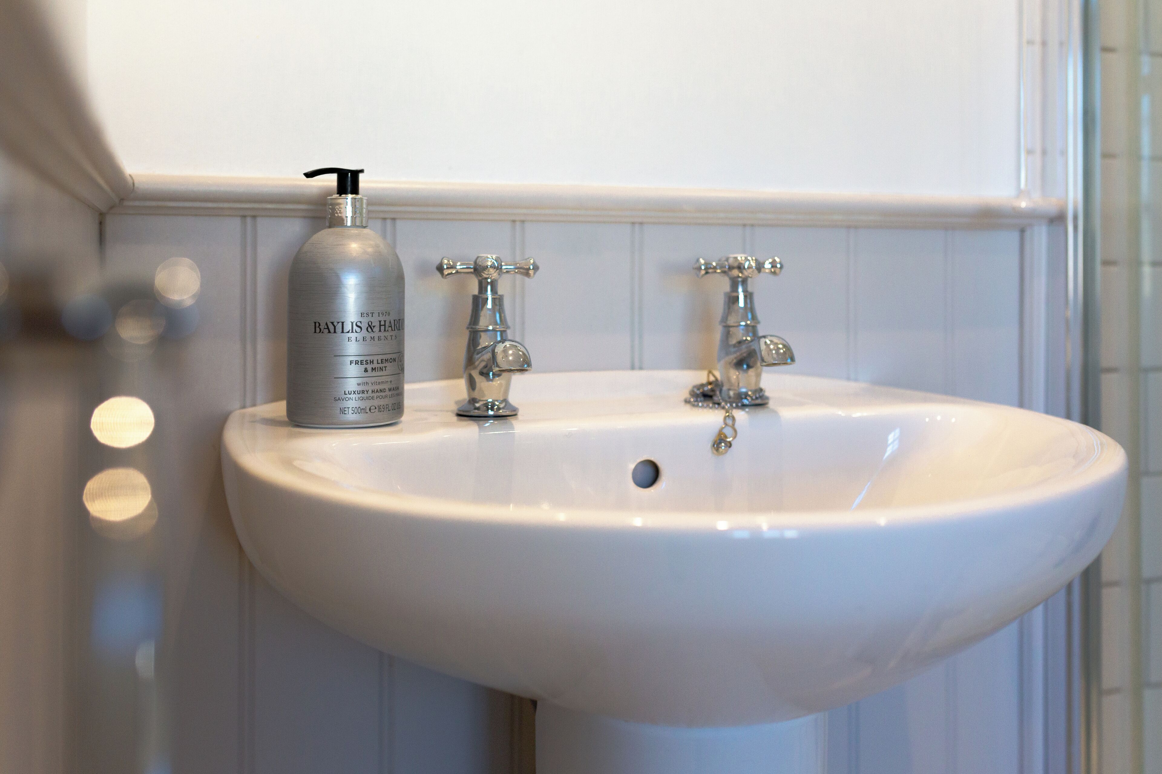 A bathroom sink with a Baylis & Hensley hand soap dispenser, two chrome faucets, and a white tiled wall.