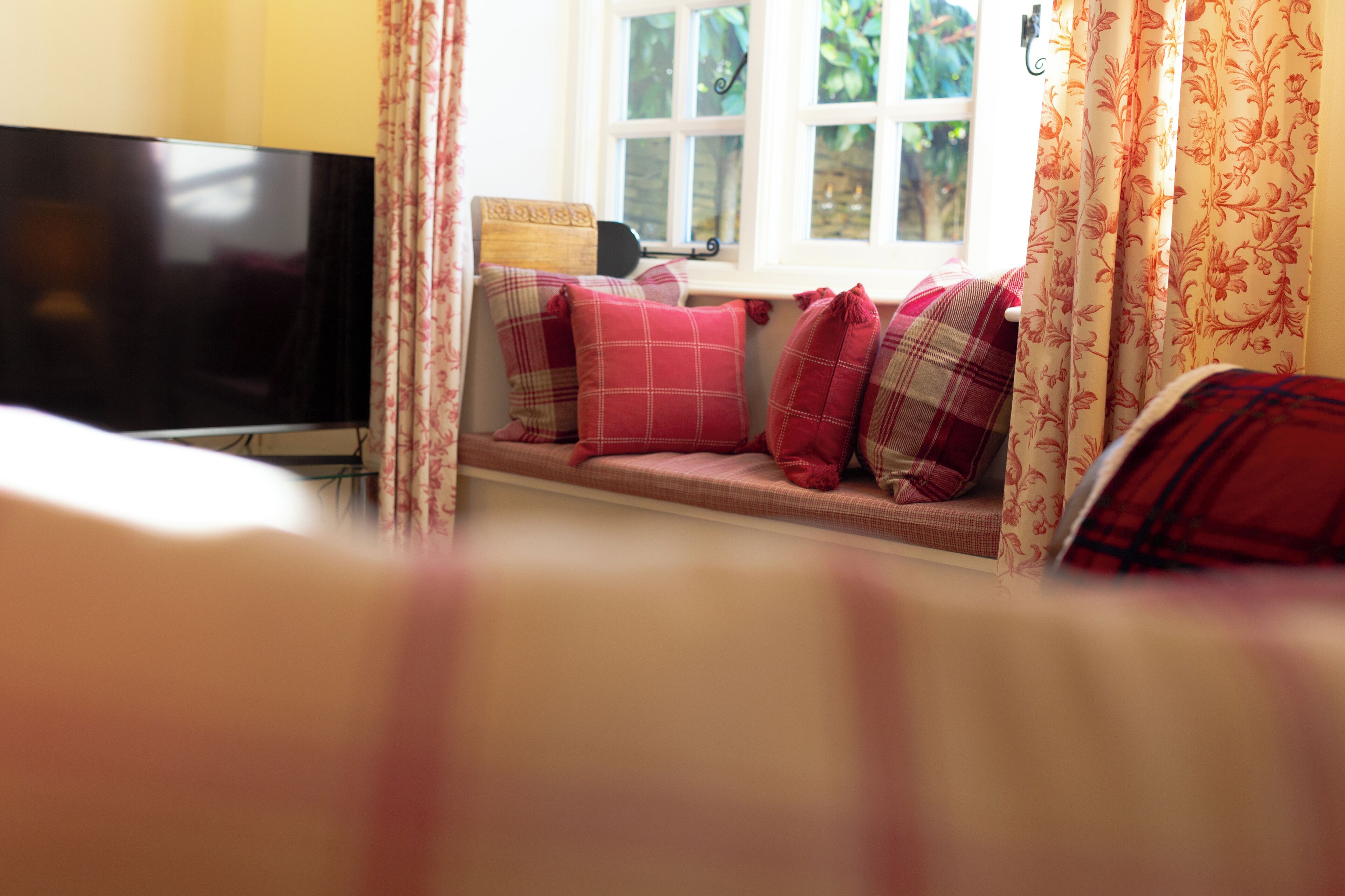 A cozy room with a television on a stand, a window with curtains, and a bench with pillows.