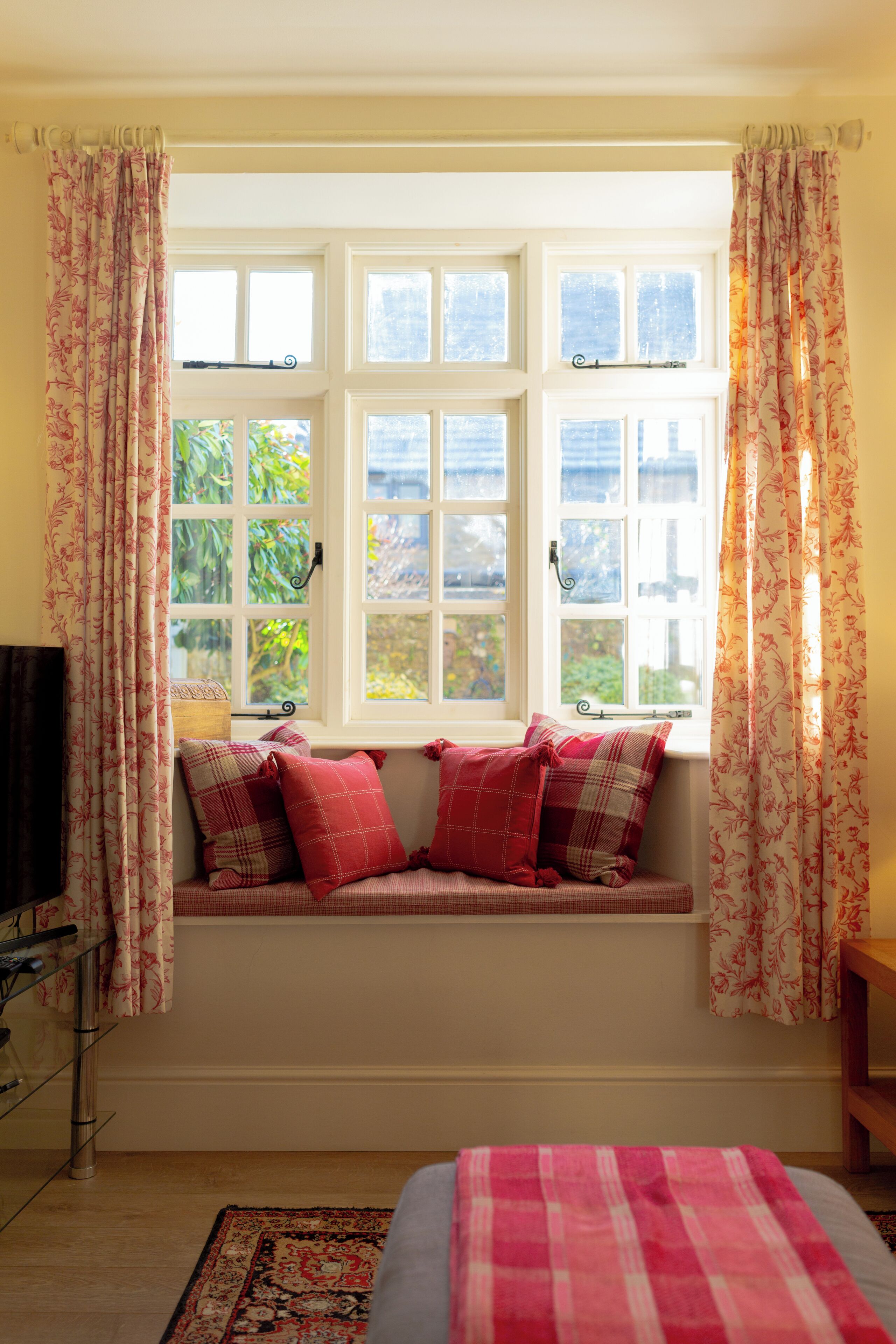 A room with a bay window, a bed with a pink and white checkered bedspread, and a red and white patterned rug.