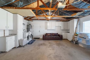 A room with a high ceiling, exposed wooden beams, white cabinets, a washing machine, a brown leather couch, and a window with blinds. - Blue Coyote Whole House (Yucca Valley)