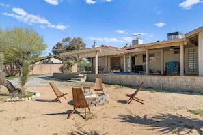 A covered outdoor seating area with a fire pit, lounge chairs, and a patio with a view of a house.