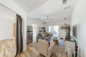 A spacious living area with a sofa, a coffee table, and a dining table. There is a kitchen area with a refrigerator and a microwave. The room has a ceiling fan and large windows. - Blue Coyote Whole House (Yucca Valley)