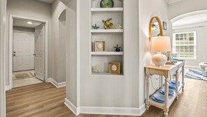 Un pasillo con una puerta blanca, suelo de madera, una pared blanca con estante y una mesa con lámpara.