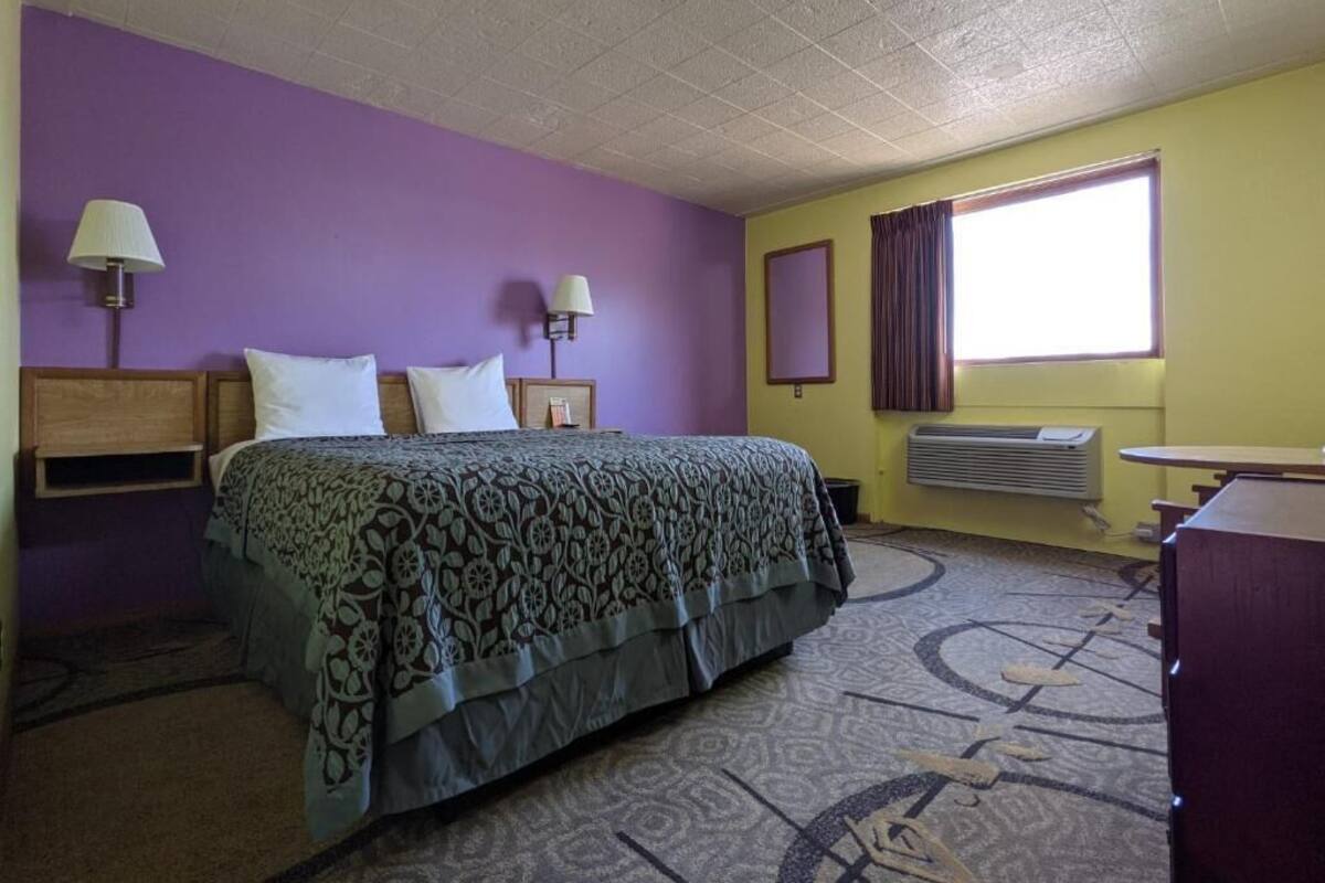 A hotel room with a bed, two bedside lamps, a window with curtains, an air conditioning unit, and a desk with a chair.