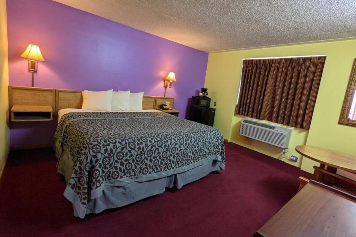 A hotel room with a bed, two bedside lamps, a heater, a table, and a window with curtains.