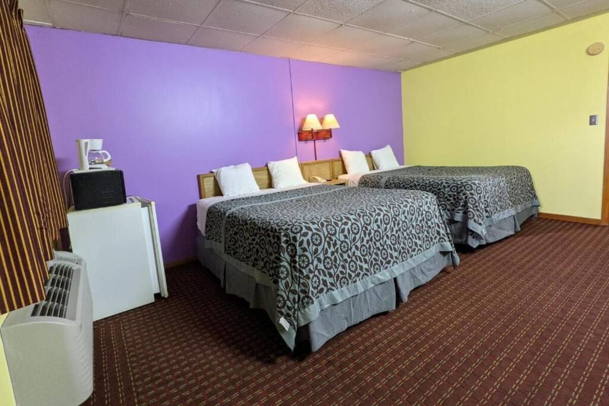 A hotel room with two beds, a desk, a lamp, and a radiator.