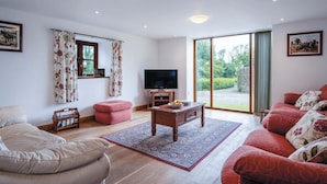 Una sala de estar moderna con sofá, reposapiés y una mesa de centro. Hay un televisor y una ventana con cortinas.