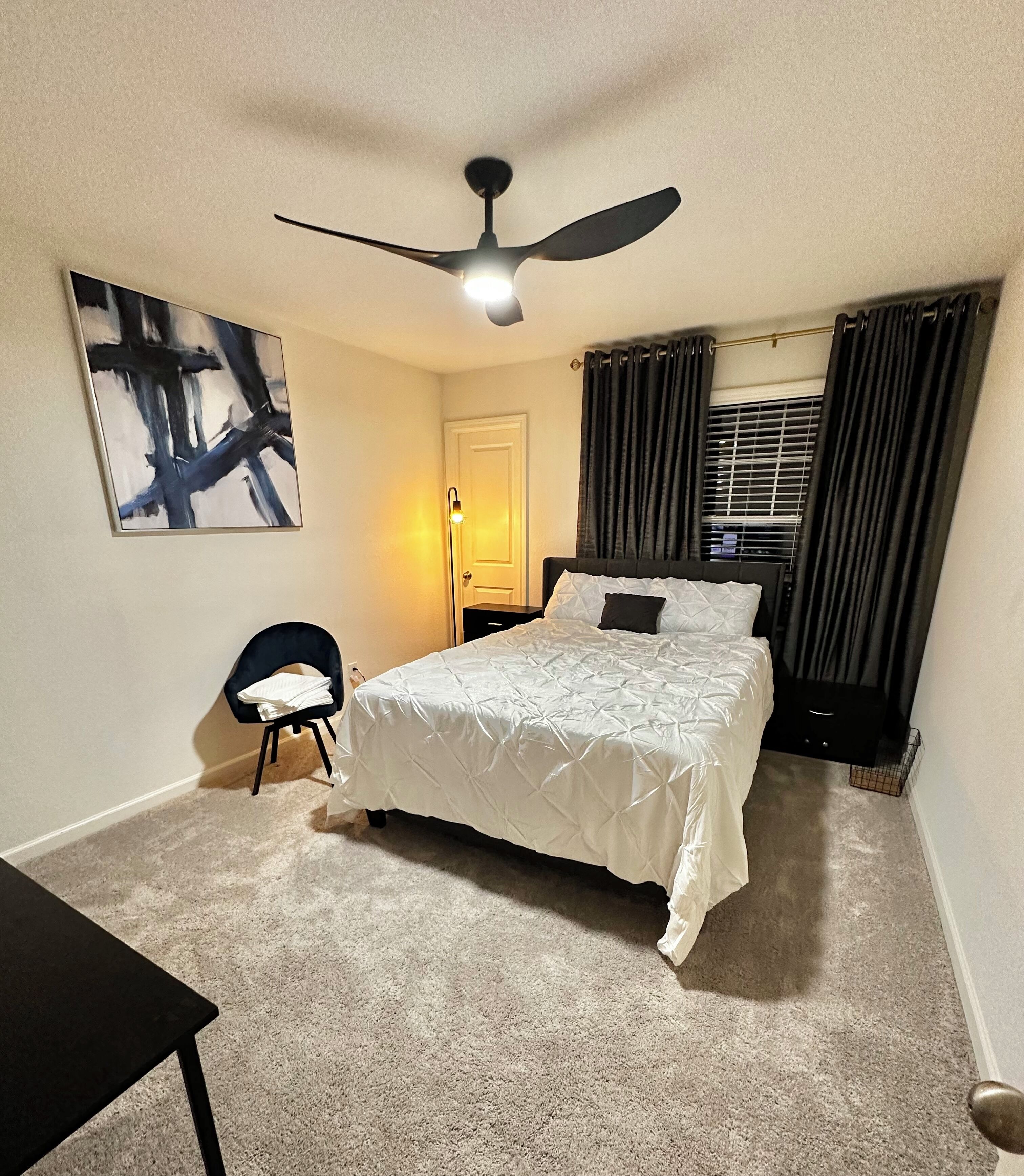 A bedroom with a bed, a desk, a chair, a ceiling fan, and a window with curtains.