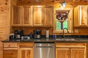 A wooden cabin kitchen with built-in cabinets, a stainless steel dishwasher, a coffee maker, and a window with a view of greenery. - Moonlight Ridge Lodge - Hocking Hills (Logan)