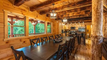 A wooden cabin interior with a dining area, kitchen, and large windows offering a view of the forest.