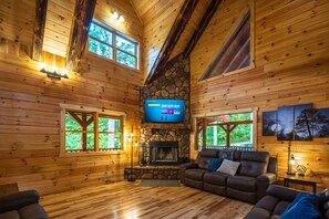 A cozy cabin interior with wooden walls, a stone fireplace, a flat-screen TV, and a comfortable seating area.