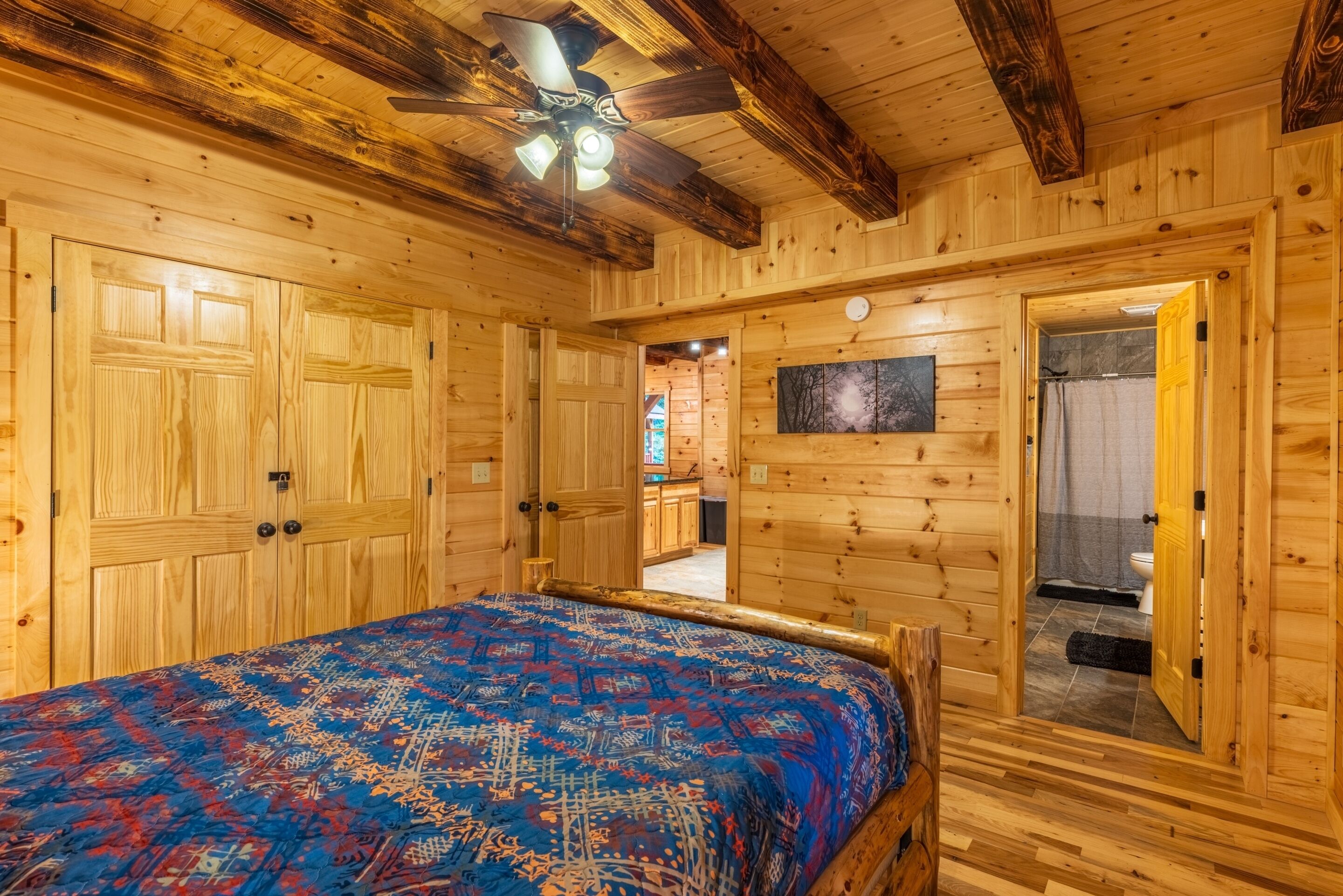 A wooden cabin bedroom with a bed, a ceiling fan, and a bathroom visible through an open door.