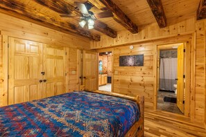 A wooden cabin bedroom with a bed, a ceiling fan, and a bathroom visible through an open door.