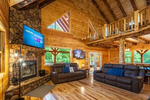 A wooden cabin interior with a stone fireplace, two leather sofas, a TV mounted on the wall, and a balcony. - Moonlight Ridge Lodge - Hocking Hills (Logan)