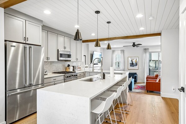 A modern kitchen with stainless steel appliances, a large island, pendant lighting, and a view to a living area with a sofa and a window.