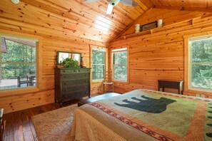 Un dormitorio tipo cabaña de madera con paredes de madera, una cama grande con colcha estampada, un aparador y una ventana con vistas a una terraza.