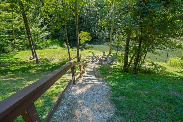 Ein Kiesweg führt zu einer Picknickfläche mit Bänken und einer Feuerstelle, umgeben von Bäumen und einem Gewässer.