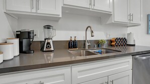 Eine moderne Küche mit weißen Schränken, einer Edelstahl-Spüle, einer Kaffeemaschine, einem Mixer und einem Weinkühler.