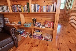 Una habitación acogedora con un sillón de cuero, estantes de madera llenos de libros y juegos de mesa, y una cesta de mimbre en el suelo.