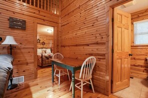 Interior de una cabaña de madera con una mesa de comedor y sillas, una cama con lámpara, y una puerta que da a otra habitación.