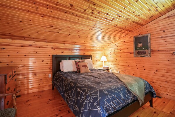 Un'accogliente camera da letto di una baita con soffitto in legno, un letto con copriletto decorato, un comodino con lampada e un quadro appeso al muro.