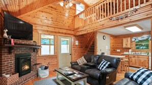 A cozy log cabin interior with a fireplace, leather sofa, and a kitchen area. - Hidden Falls Retreat | Hocking Hills, Waterfall (Rockbridge)