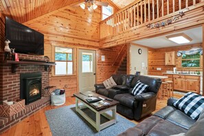 A cozy log cabin interior with a fireplace, leather sofa, and a kitchen area.