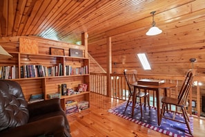 Accogliente interno di una baita di legno con una poltrona in pelle, mensole in legno piene di libri, un tavolo da pranzo con sedie e vista sulla natura attraverso una finestra.
