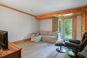 Una habitación acogedora con techo de madera, pared de ladrillo blanco, un sofá, un sillón reclinable y un televisor sobre un mueble de madera.